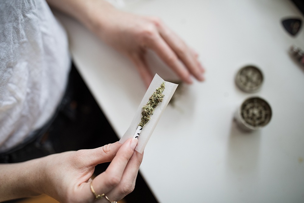 A teen holds rolling papers with cannabis in them and on the table