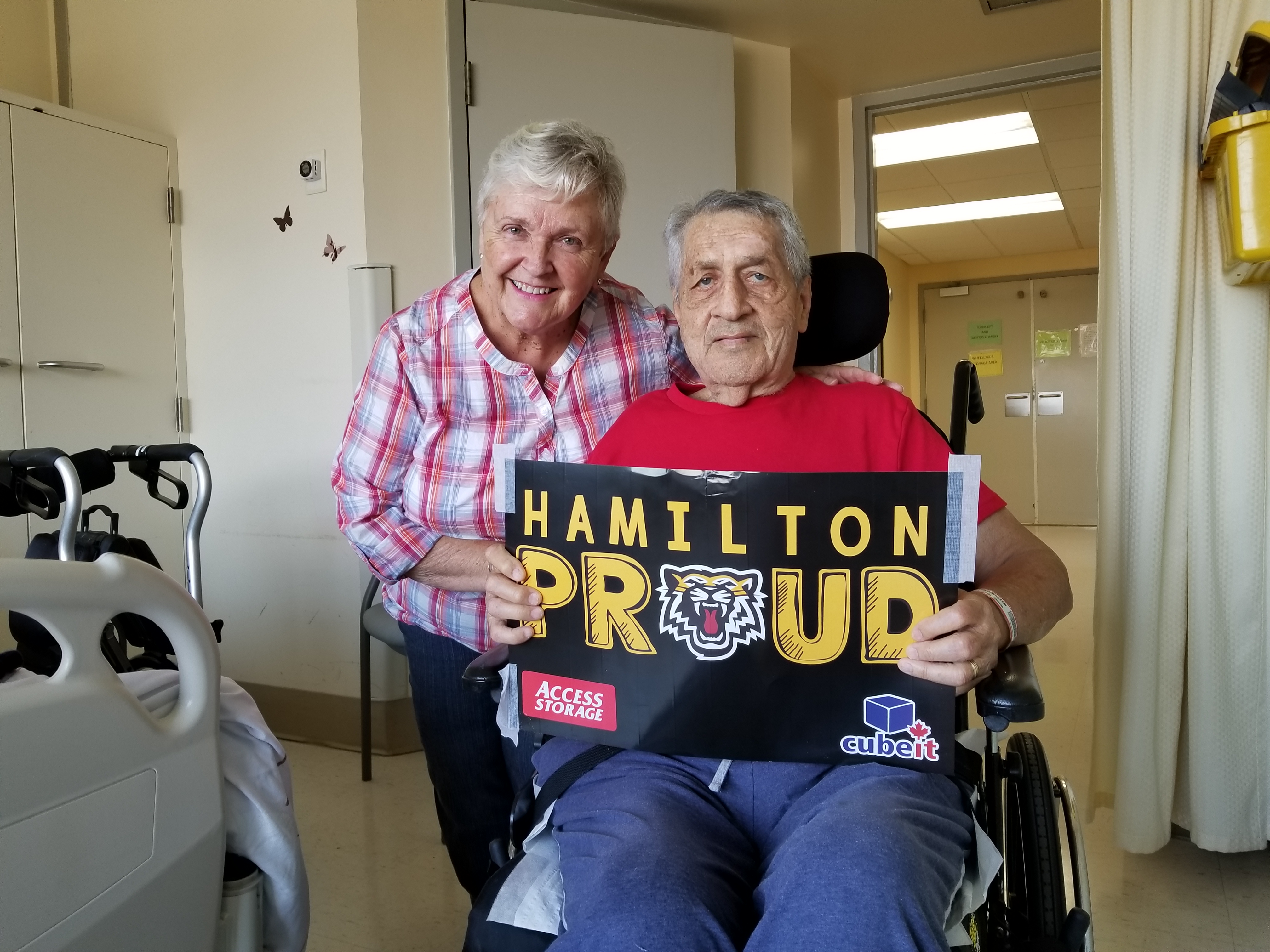 Kathy poses with her husband Bill, holding a Hamilton Proud sign