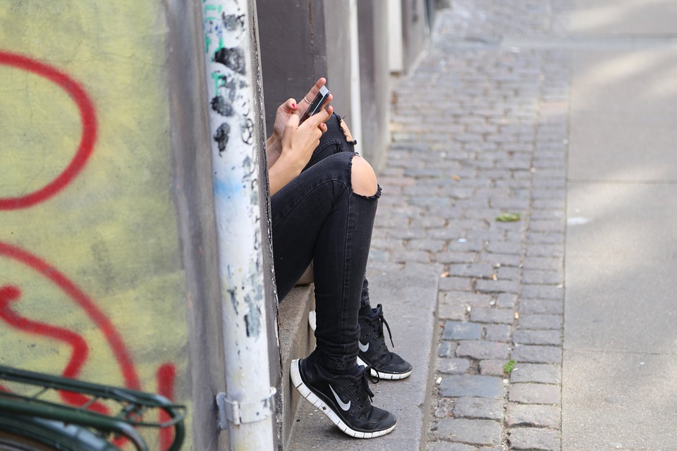 A teenage girs' knees in ripped jeans are peeking out from a doorway where she is sitting, texting on her phone