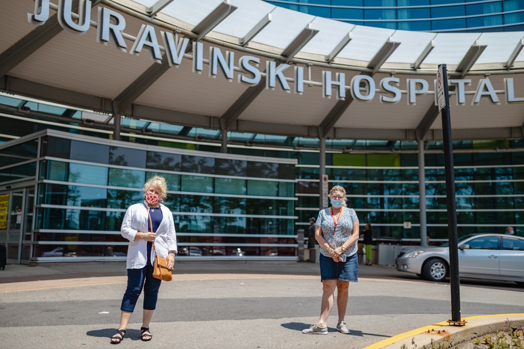Family caregiver Jeanette Arsenault and social worker Victoria Gibbins stand outside the Juravinski Hospital main entrance. The hospital sign is visible above their heads.