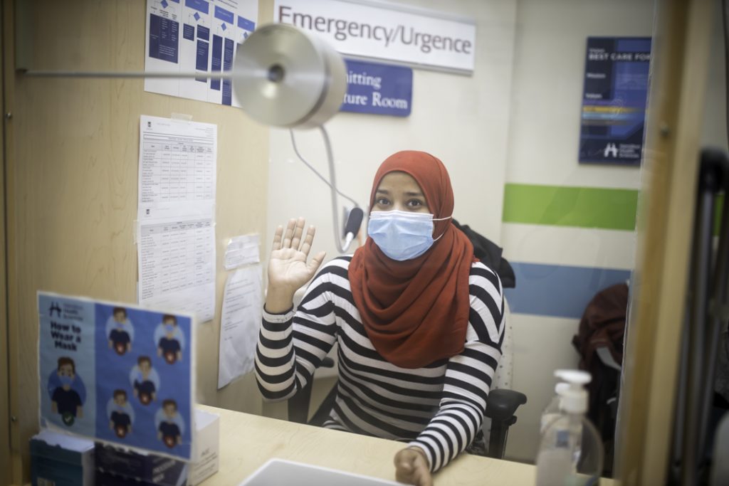 A woman sits behind plexiglass wearing a medical mask