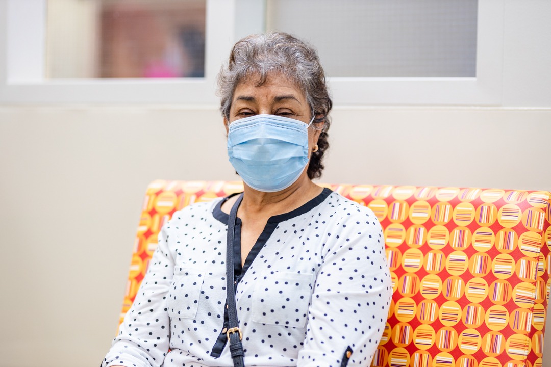 A woman wears a medical mask and sits in a hospital waiting room on a brightly-coloured chair
