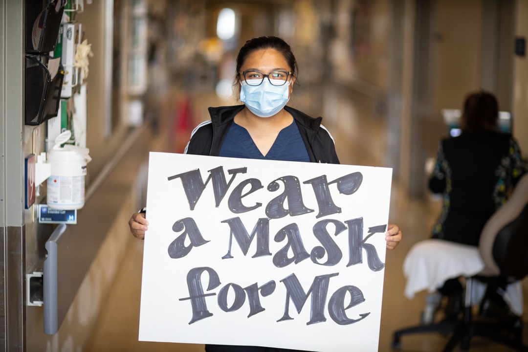 A health care worker wearing a mask holds a large sign reading "wear a mask for me"