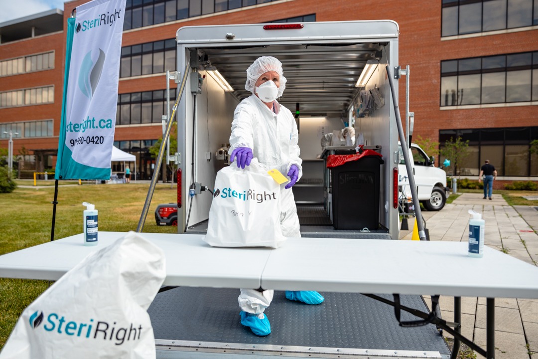 A person wearing a white uniform and N95 mask hands a white back out of the back of a truck onto a table