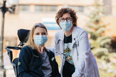 Two women pose together wearing medical masks. One woman is using a wheelchair.
