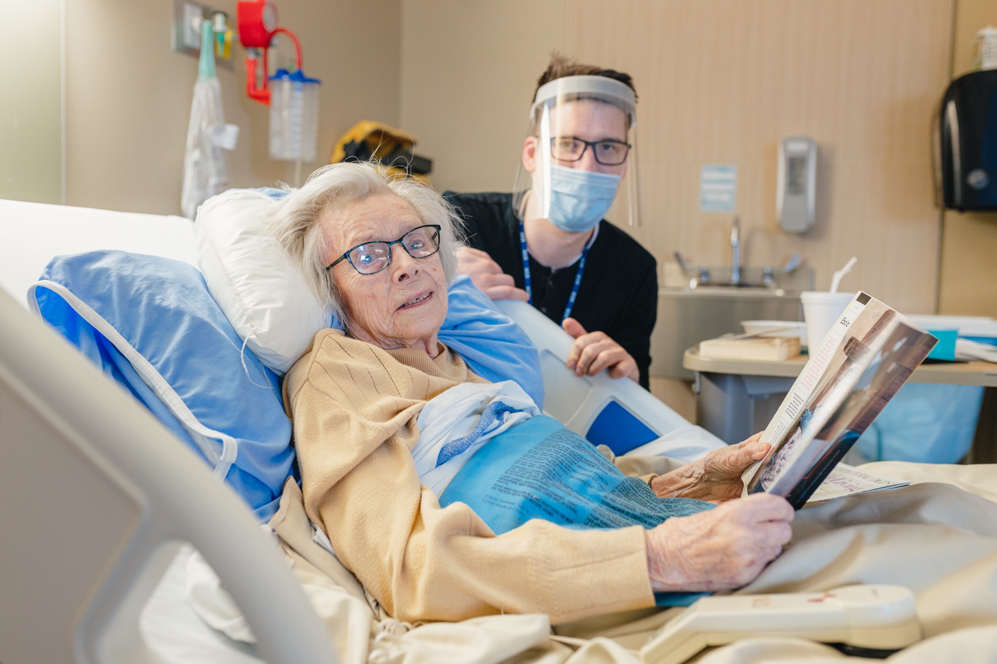 Malise Lynch smiles at the camera from her hospital bed. Elder life specialist Chris Gabor sits beside her wearing personal protective equipment.