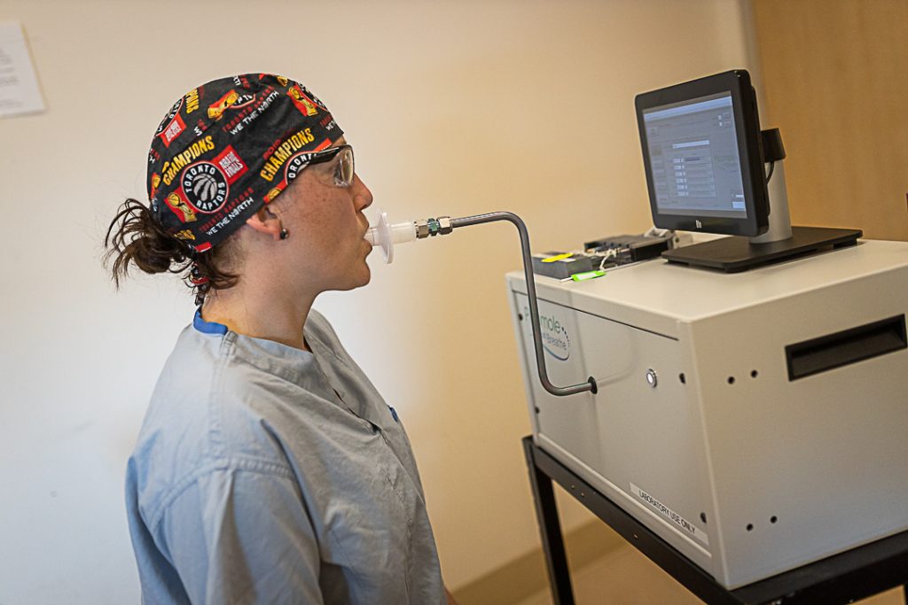 A nurse wearing a Raptors scrub cap breathes into a straw-like tube connected to a machine and computer.
