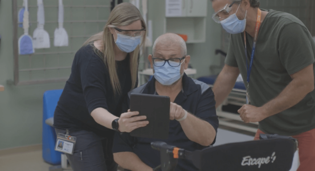 Two health care workers assist a patient in using Epic on a tablet.