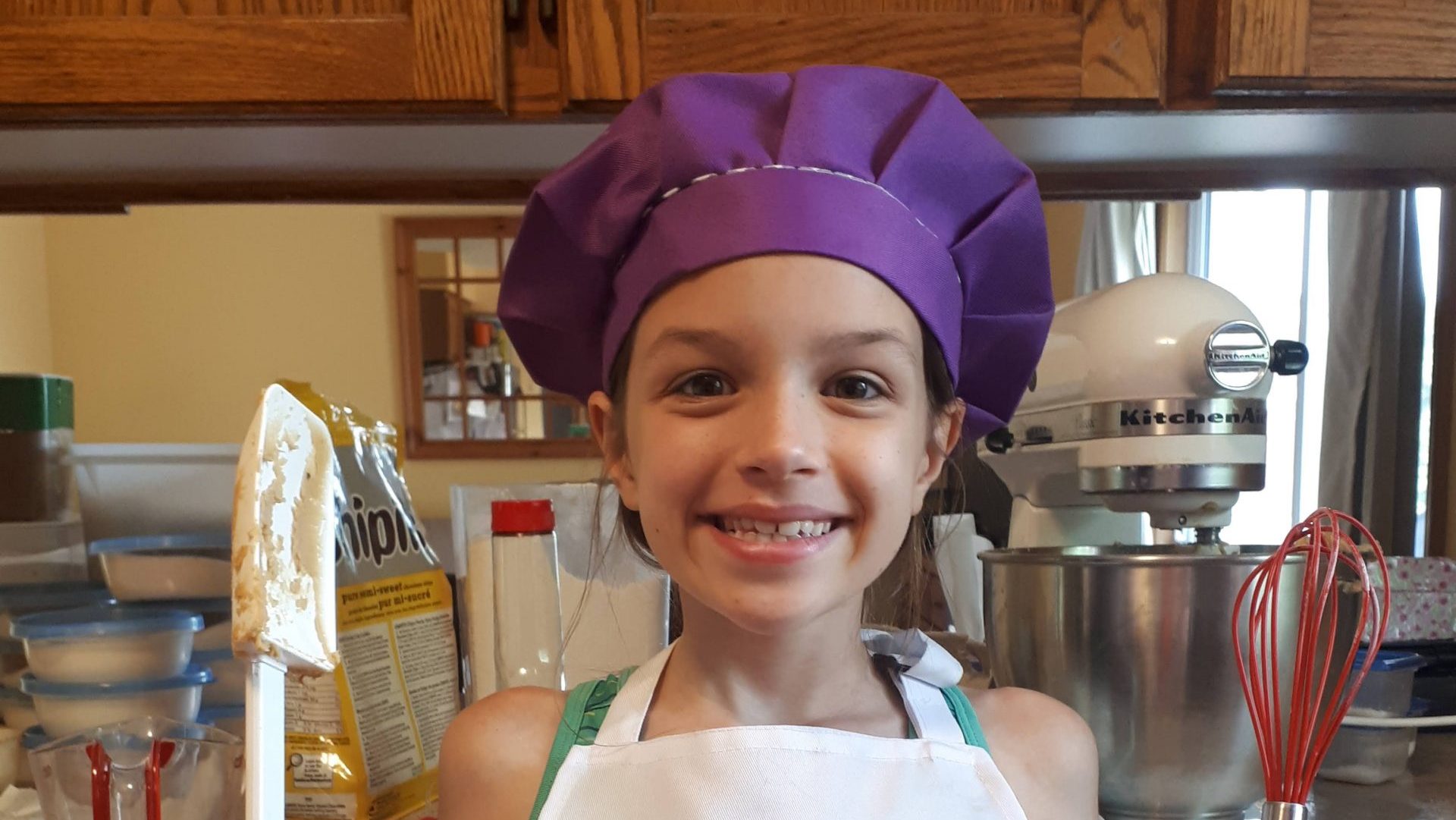 Alyssa Lodge standing in her kitchen wearing a purple chef's hat, white apron with her name displayed, holding kitchen tools in each hand
