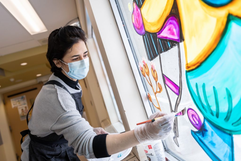 A woman wearing a surgical mask and an artist's apron paints a window with bright colours