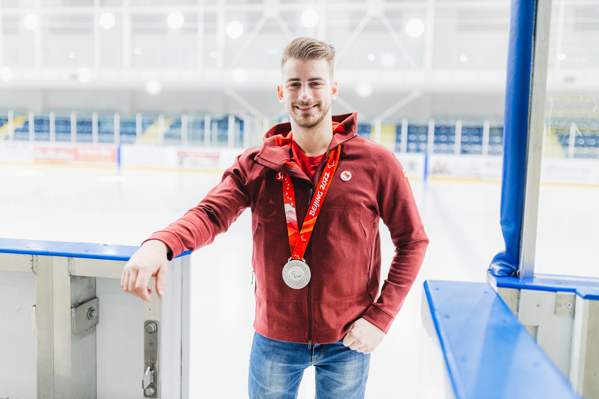 Young man stands at the entry way of an indoor ice pad wearing a red jacket and silver medal around his neck