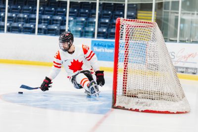 A sledge hockey player wearing a Team Canada white jersey with red maple leaf shoots at the net