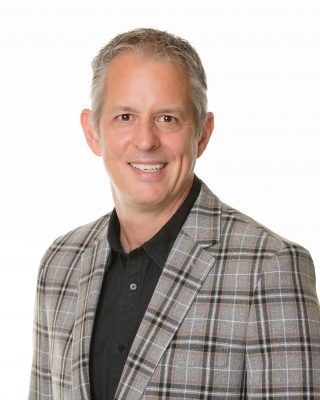 Head and shoulders photo of Dr. Devin Peterson, wearing a black collared shirt and grey checked sports coat