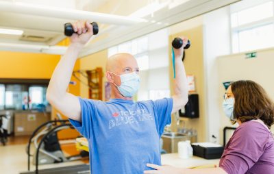 A man, wearing a paper mask, holds to small hand weights above his head. He is wearing a light blue T-shirt and working out at the HHS Regional Rehabilitation Centre fitness centre.