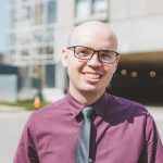 Eric Ricker, bald, black frame glasses, standing outside McMaster University Medical Centre, wearing a purple button down shirt and black tie