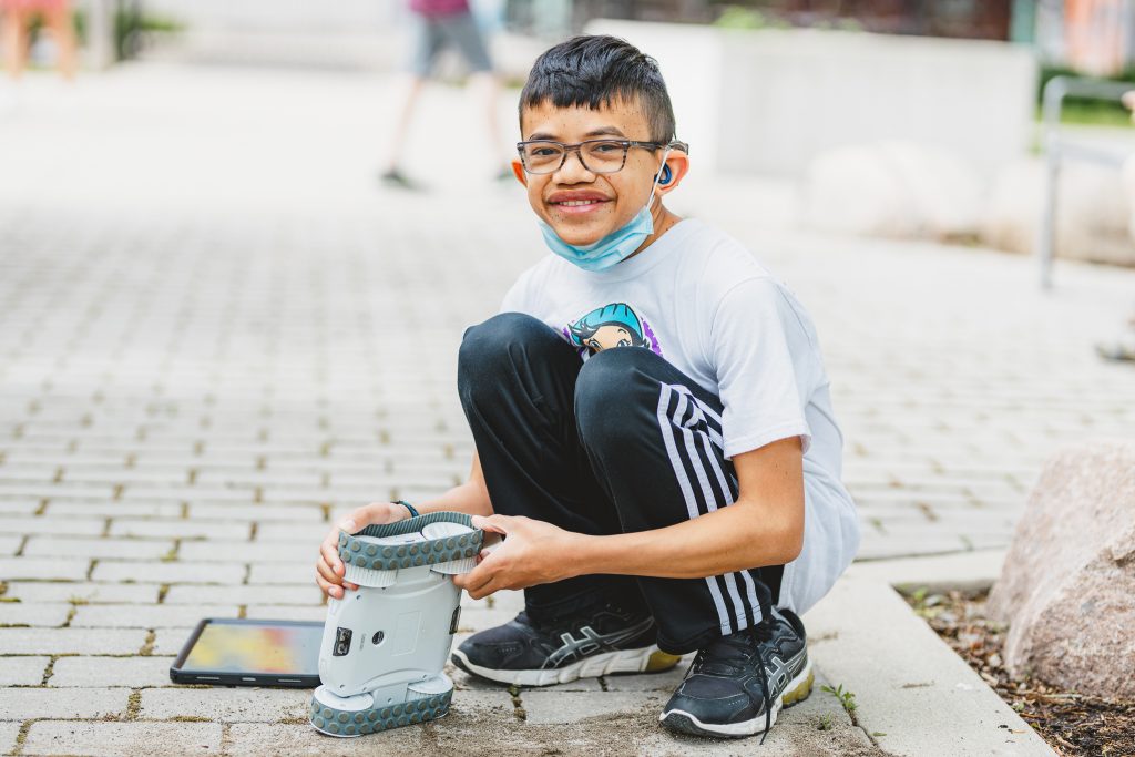 A boy crouches on the ground, playing with a robotic toy outdoors