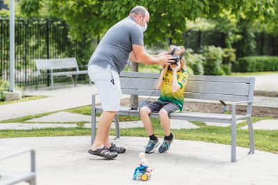 A man helps a boy adjust virtual reality goggles. They are outdoors and the boy is sitting on a bench