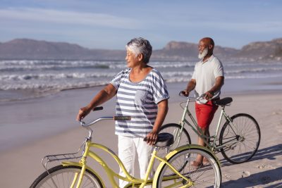 Two older adults stand on a beach beside their bicycles
