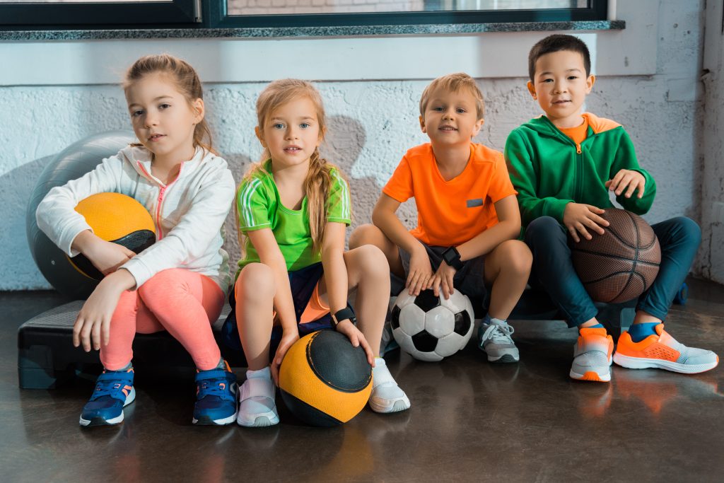Multicultural children sitting on step platforms with balls in gym