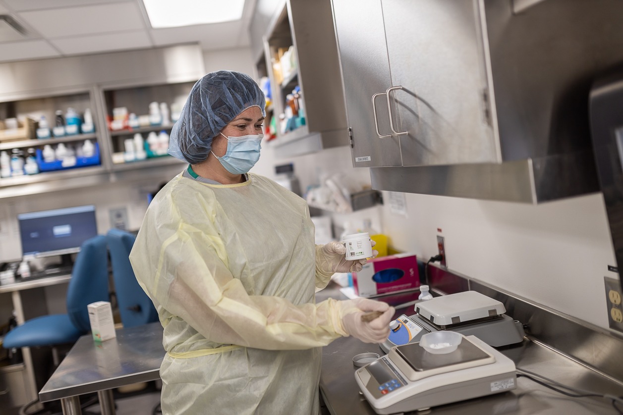A hospital worker wears a blue surgical cap, blue facemask and yellow scrubs while weighing a pharmaceutical powder.