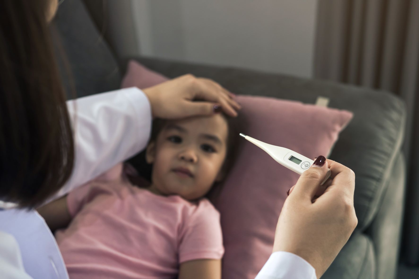 An adult is holding a thermometer in one hand and has their other hand on a child's forehead. The child is about three years old and is lying in bed on a pink pillow.