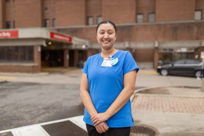 A young woman wearing a blue shirt and hospital ID tags stands outdoors in front of the Hamilton General Hospital emergency department entrance.