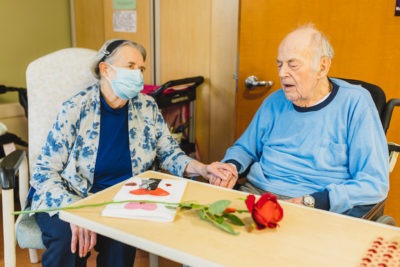 An older woman hold hands with her husband at St. Peter's Hospital. A rose and homemade Valentine's Day card rest on a table