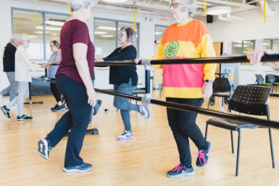 Older adults, in a gym, practise balance exercises while holding on to a ballet bar