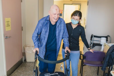 A clinical extern supports an older patient, using a walker to walk down a hospital hallway