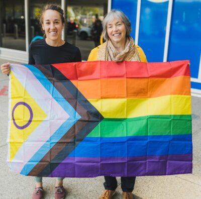 Stephanie Tibelius and Monica Bennett are incoming co-chairs of Hamilton Health Sciences new Pride Employee Affinity Group. They're holding a flag representing the Two-Spirit and LGBTQIA+ community.