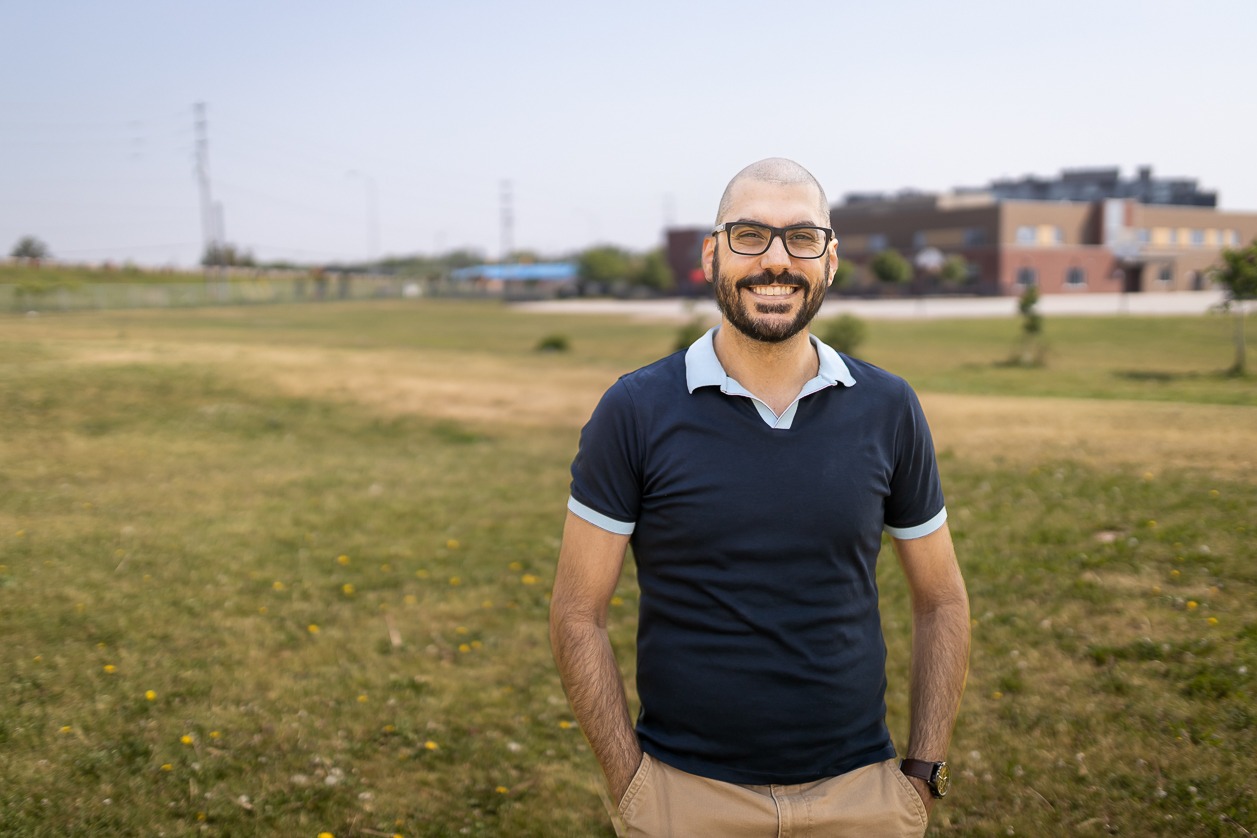 Alex Seixiero stands in a park with a grassy field behind him. He is a young man wearing glasses and a dark collared t-shirt.