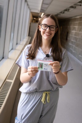 A young woman wearing hospital scrubs holds a staff ID badge.