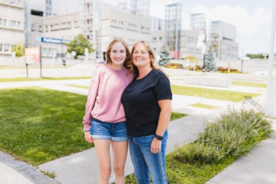 A teenager and her mother stand with their arms around each other. The McMaster Children's Hospital is in the background.