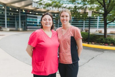 Gillian Crawford and Francesca Schaaf are standing together outside the main entrance of Juravinski Hospital. They are both wearing pink scrubs and smiling. 