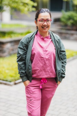 Jacqueline Mallari is standing outside wearing pink scrubs, glasses and an olive green jacket. Her hair is tied in a ponytail and she is smiling at the camera with her hands in her pockets. 