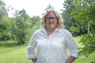 Darlene Creed is standing outside wearing a white button-down shirt and glasses. She is smiling at the camera and her hands are in her pockets. 