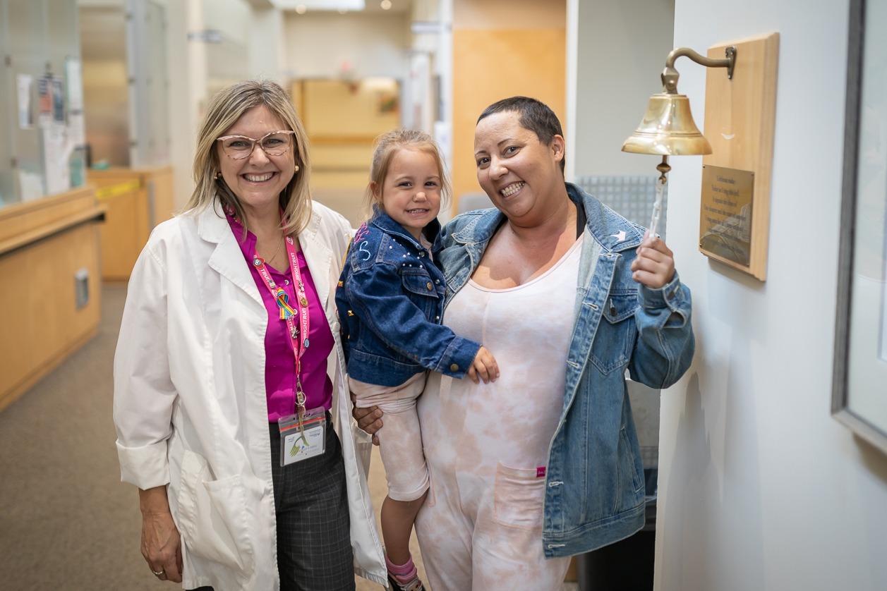 A breast cancer patient rings a bell to celebrate finishing radiation therapy. She holds her young child and stands beside a nurse.