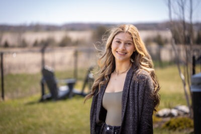 Portrait of teenager Katie Mantione smiling outdoors with a blurred background. Katie has long blonde hair and is wearing a grey shirt and grey sweater.