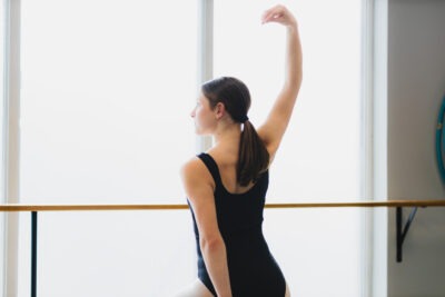 A teen girl in a black leotard pauses in the ballet studio. She is using her arms to illustrate a straight back.
