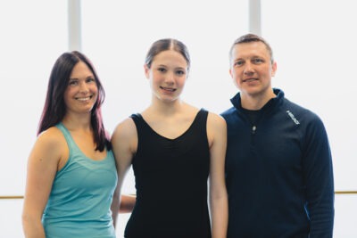 Elizabeth stands between two adults in the dance studio. They are all smiling.