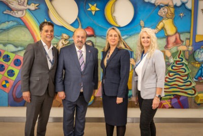 Four adults in suits stand in front of a painted mural in a children's hospital. It is a posed photo.