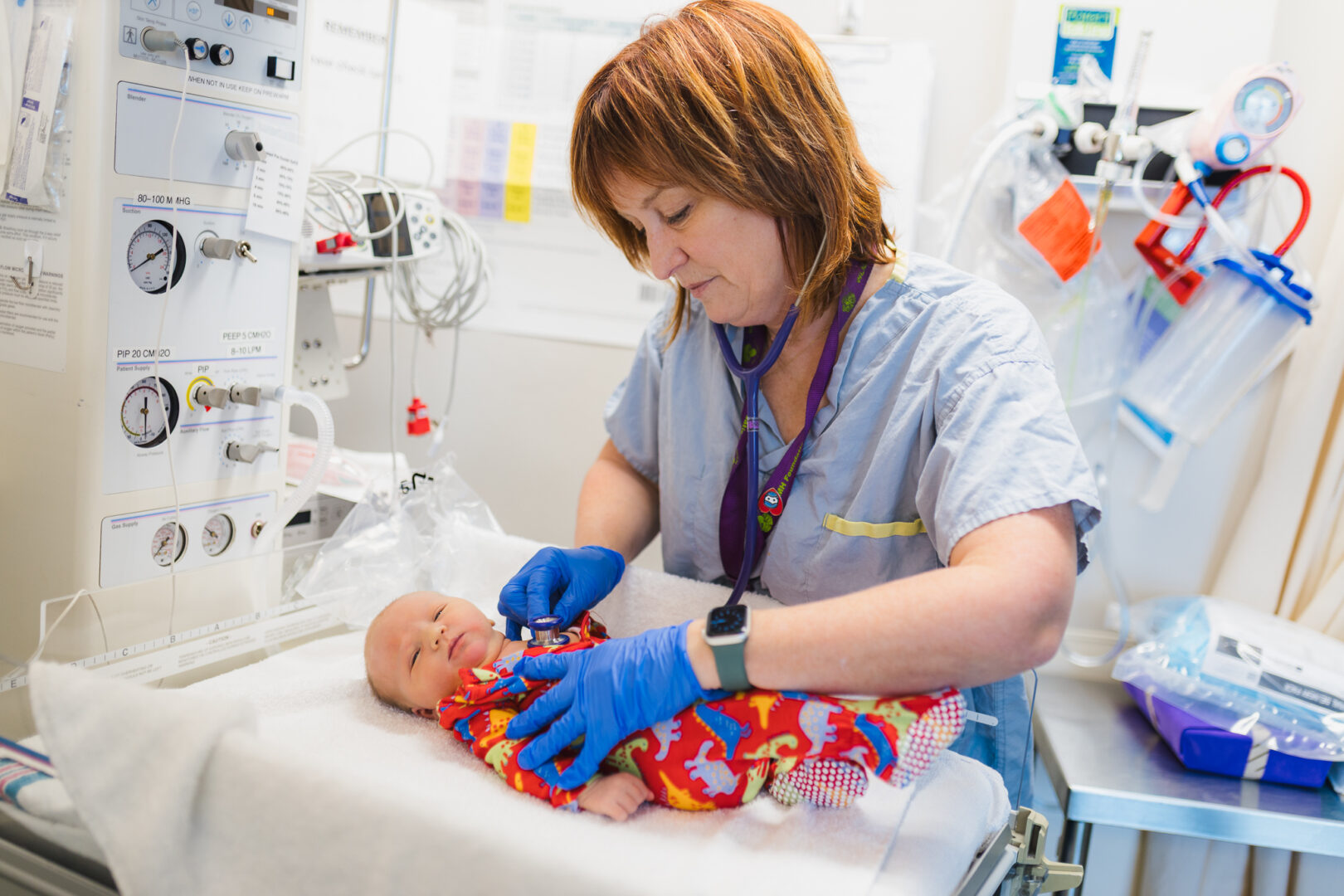A midwife supports a tiny baby on a warming table at WLMH. The baby is wearing a red onesie.
