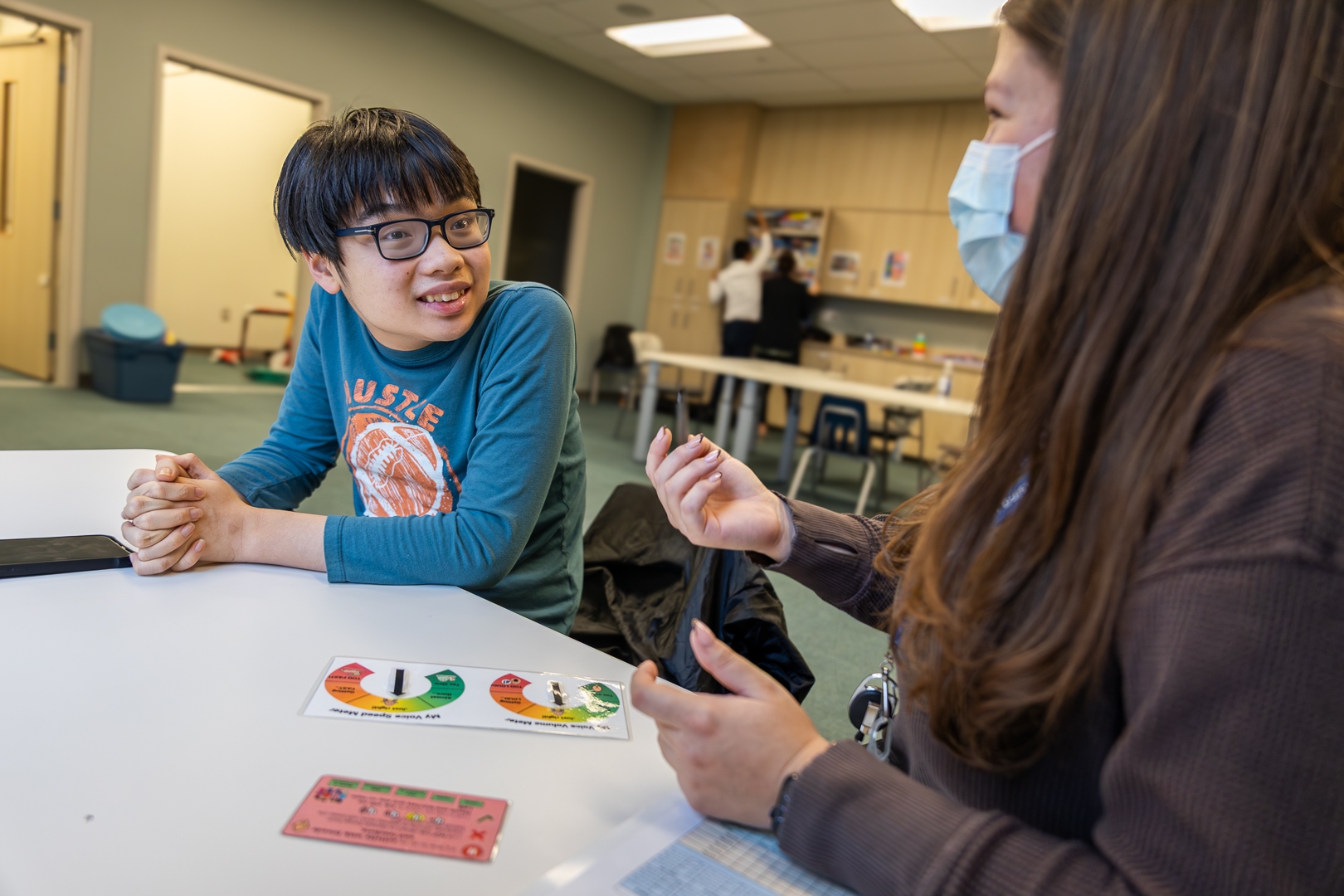 A teenager with glasses smiles while listening to a health care worker in a mask.