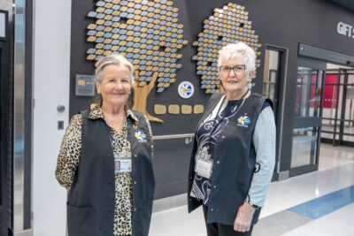 Two women in matching vests stand in front of a wall display.